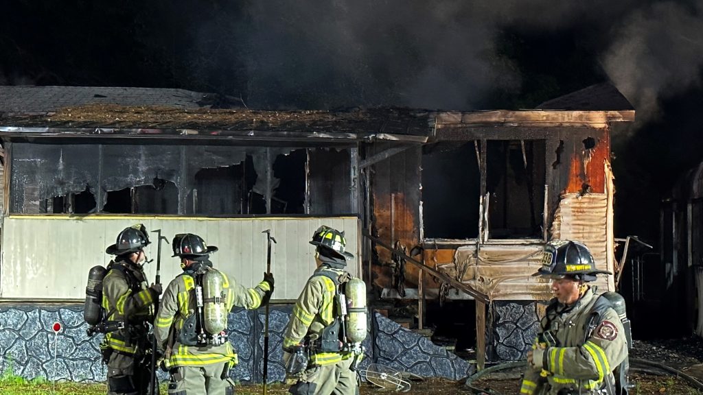 Four firefighters in full gear stand in front of a heavily damaged, burning house at night. The house is charred, with smoke rising from the roof, and the porch is partially destroyed.