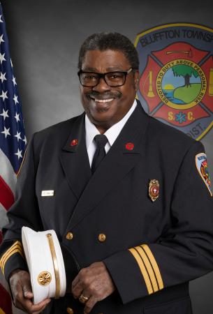 A smiling Black man wearing a formal fire department uniform stands holding a white hat. An American flag and a fire department emblem are visible in the background.