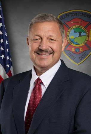A man in a dark suit, white shirt, and red tie smiles in front of an American flag and a Bluffton Township Fire District emblem.