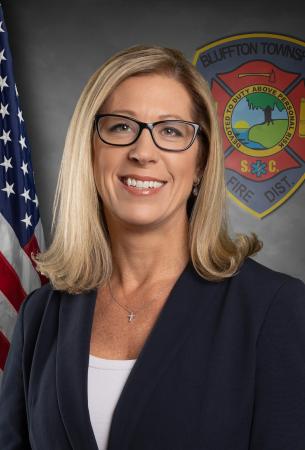 A woman with blonde hair and glasses, wearing a dark blazer and white top, smiles in front of an American flag and a Bluffton Township Fire District emblem.