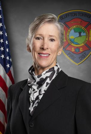 A smiling older woman with short gray hair wearing a black suit and a patterned scarf stands in front of an American flag and a fire department emblem.