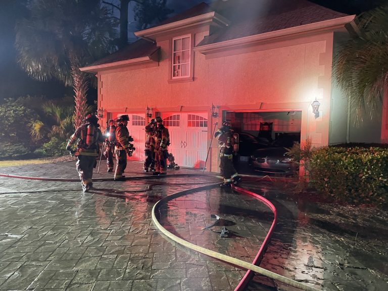 Firefighters in full gear stand outside a house garage at night, lit by red emergency lights, with a fire hose running into the open garage where a car is visible. The wet driveway reflects the lights.