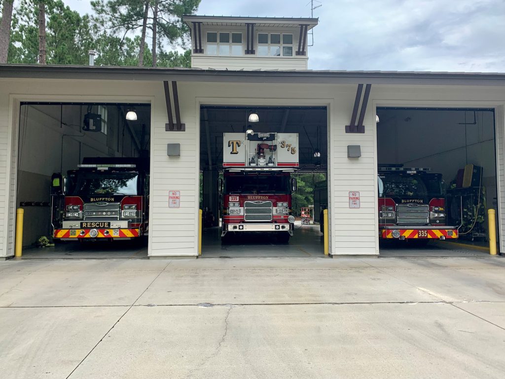 Three fire trucks are parked inside the bays of a fire station. The building is white with three large garage doors open, revealing the trucks inside. Trees can be seen in the background.