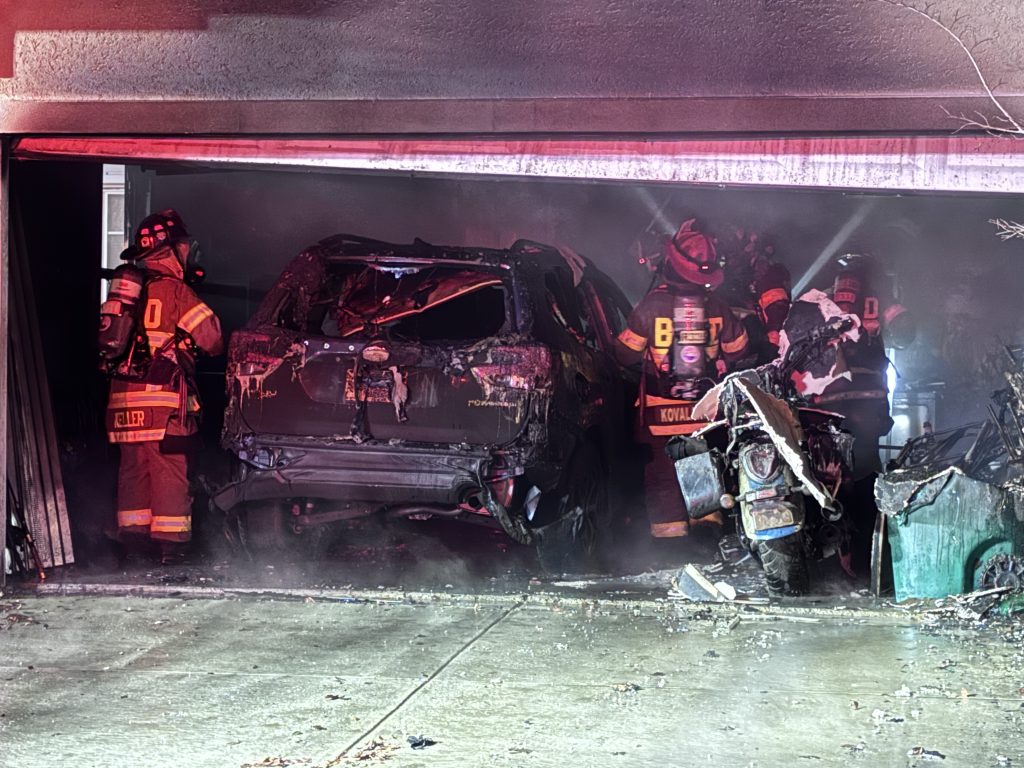Firefighters in full gear stand in a smoky garage with two heavily burned vehicles inside, working to control the aftermath of a fire. Debris is scattered on the driveway.