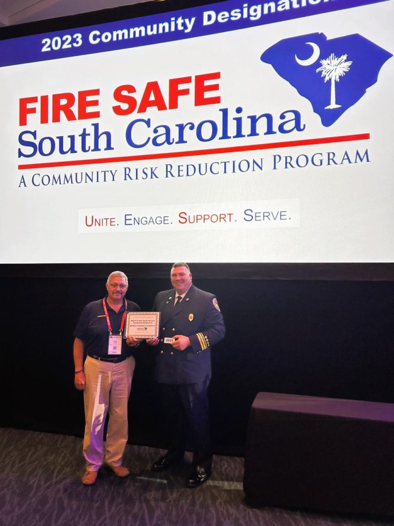 Two men stand on stage, one in a fire chief uniform holding a certificate, and the other in business casual attire. Behind them is a large screen displaying "Fire Safe South Carolina" and the program's mission statement.