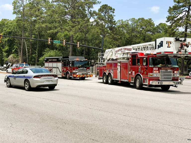 A police car, a fire truck, and a fire rescue vehicle are parked at an intersection with traffic lights. Trees and greenery are visible in the background on a sunny day.