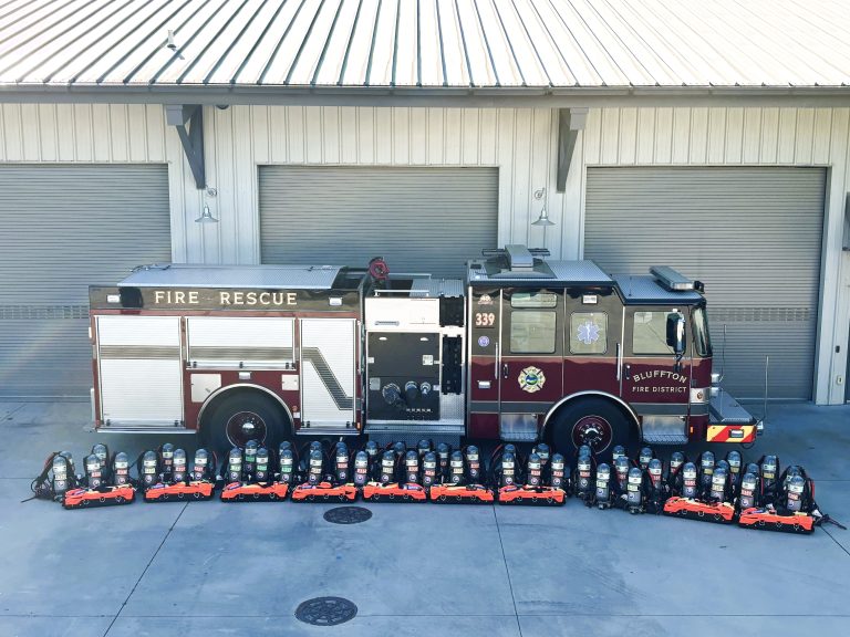 A fire engine is parked in front of a building with three closed garage doors. In front of the truck, new firefighting air packs and bright orange equipment are neatly lined up on the ground.