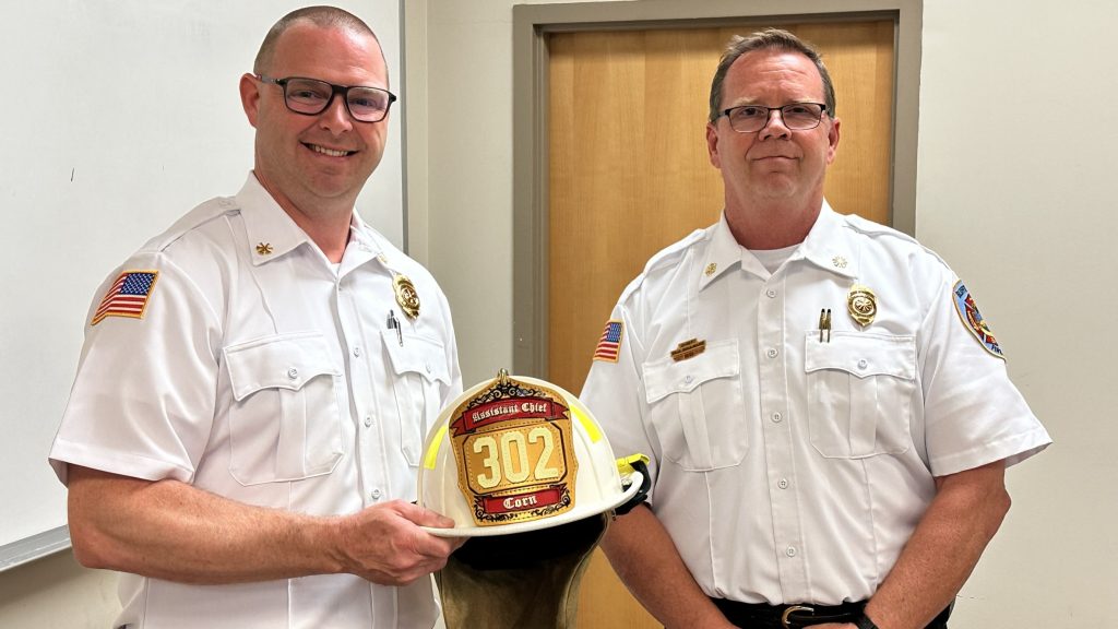 Two men in white fire department uniforms stand indoors. One holds a firefighter helmet labeled "302" with "Battalion Chief." Both men are smiling slightly and have American flag patches on their sleeves.