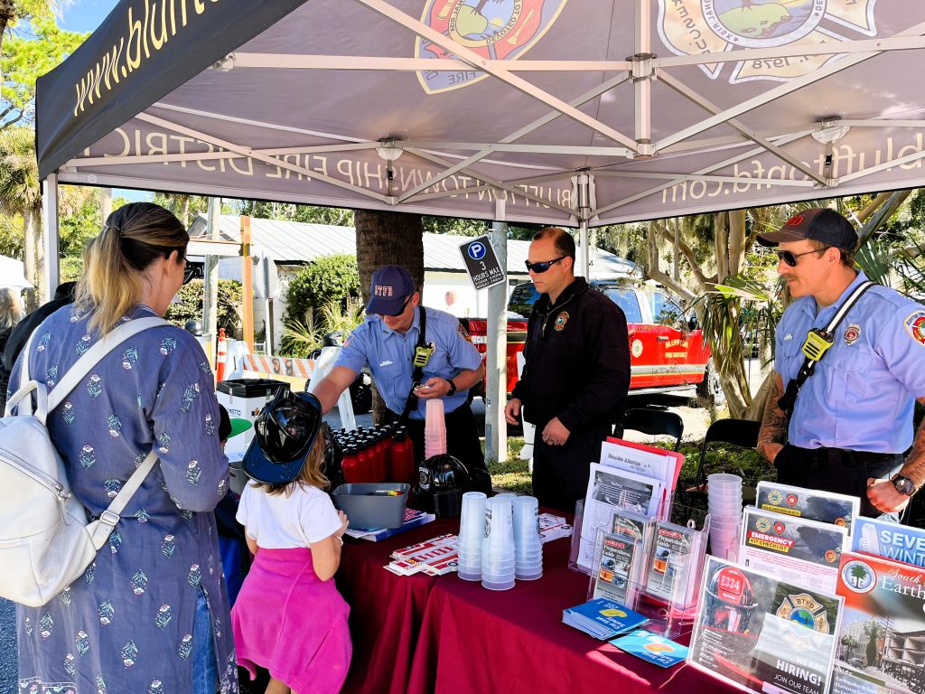 Three firefighters interact with a woman and a child at an outdoor booth under a canopy. The booth displays pamphlets, cups, and fire safety materials. The firefighters wear uniforms and sunglasses. Trees and cars are visible in the background.