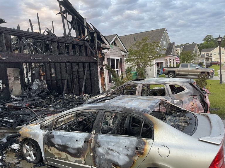 Two severely burned cars sit in front of a house with extensive fire damage; the garage and roof are charred and partially destroyed. Neighboring houses appear undamaged in the background.