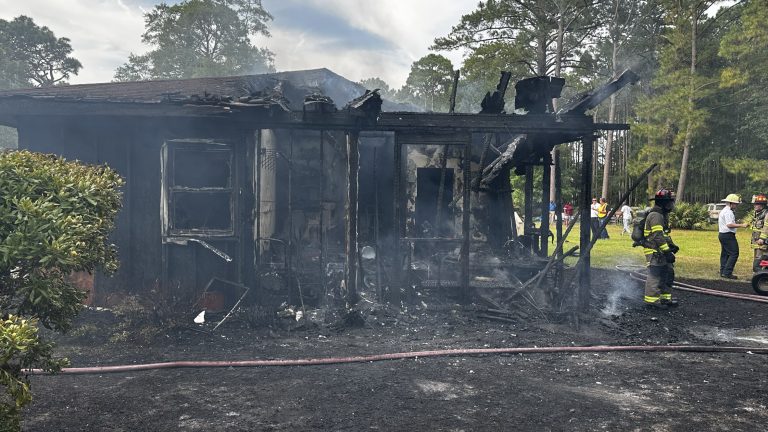 A burned house with heavy fire damage and smoke, partially collapsed roof and walls. Firefighters in gear stand nearby, with hoses on the ground and trees visible in the background.
