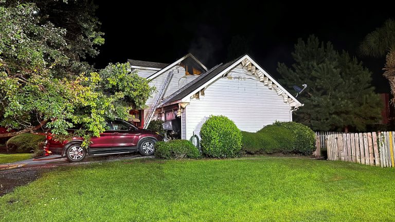 A house with fire damage is seen at night. The siding is charred near the garage, with scorch marks and smoke stains. A red SUV is parked nearby, surrounded by green bushes and a wooden fence. The yard is brightly lit.