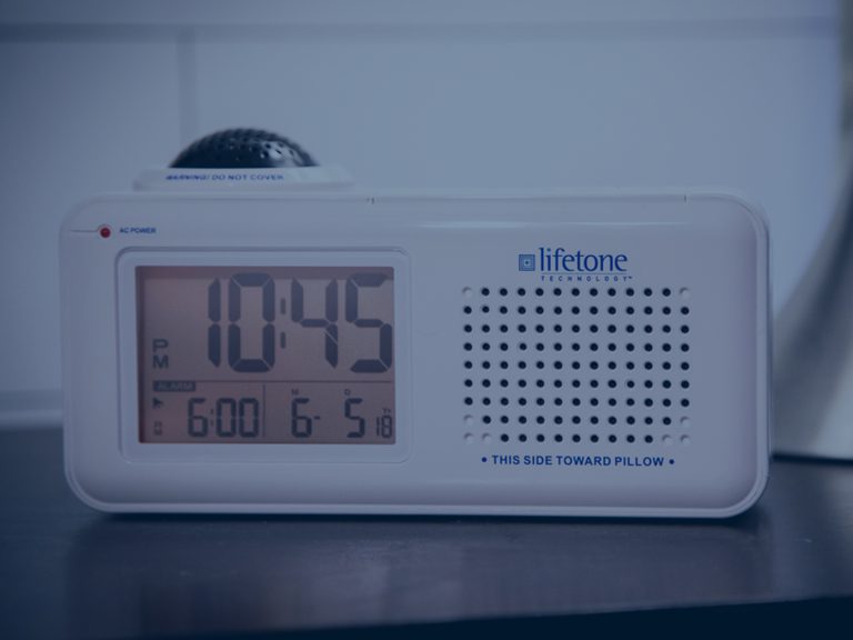 A white LIFETONE bedside alarm clock with a digital display shows the time as 10:45 and alarms set for 6:00 AM and 6:15 AM. The device is on a dark surface with a blurred background.