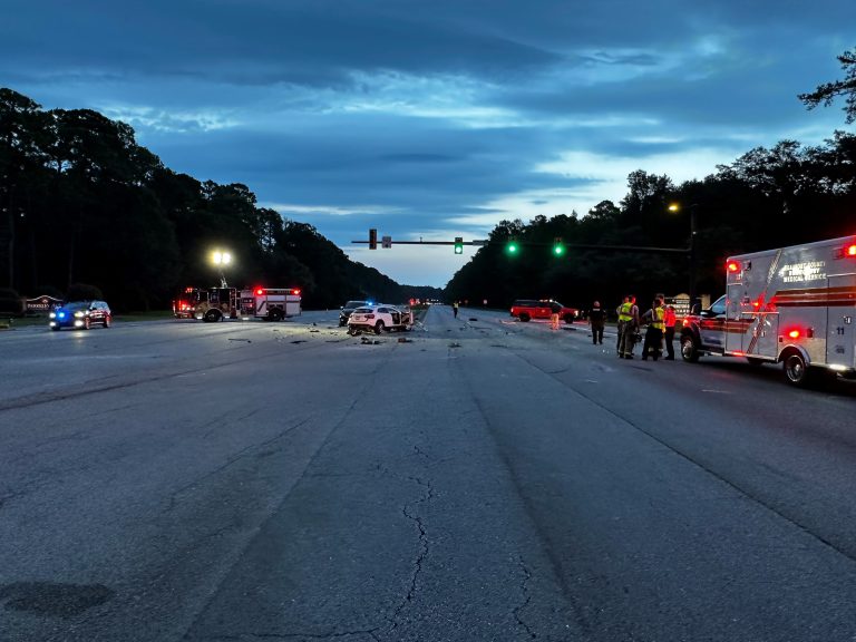 A wide road at dusk with emergency vehicles, including police cars, fire trucks, and ambulances, at the scene of an accident. Debris is scattered on the road, and several responders are standing near the vehicles.