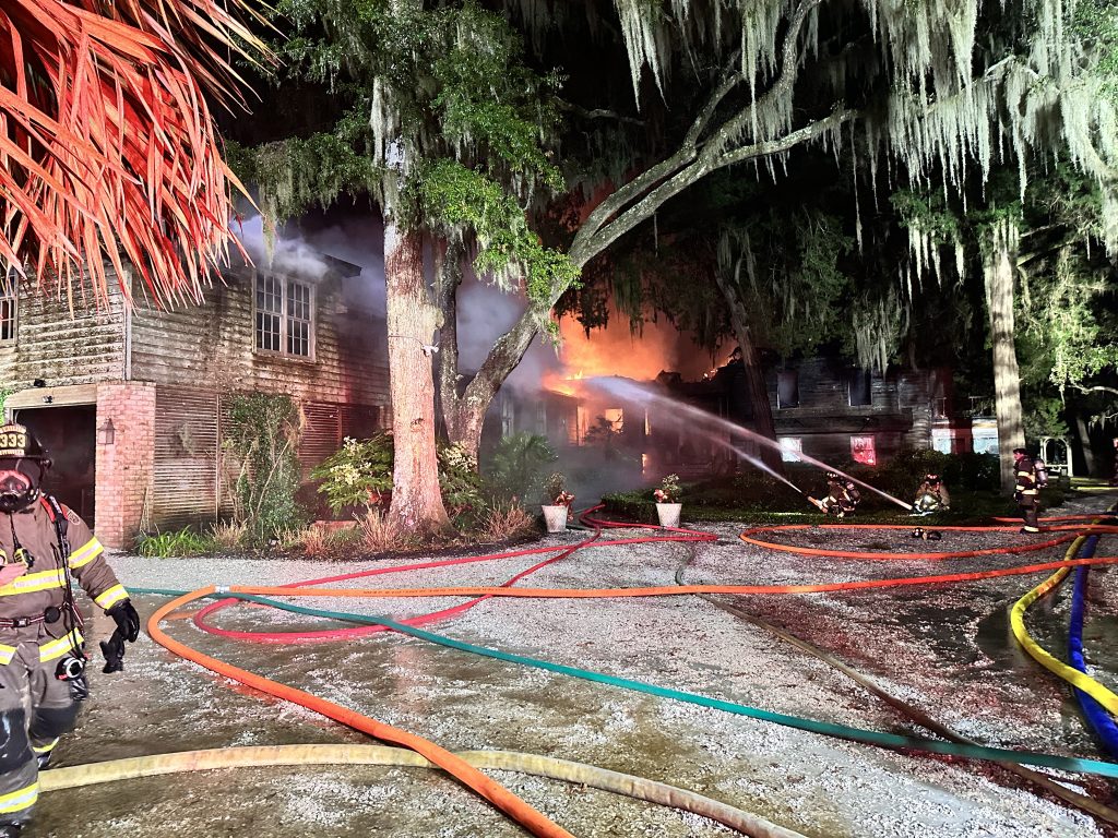 Firefighters spray water on a burning house at night, surrounded by trees and heavy smoke. Multiple fire hoses are laid across the ground, and flames are visible through the building’s windows.