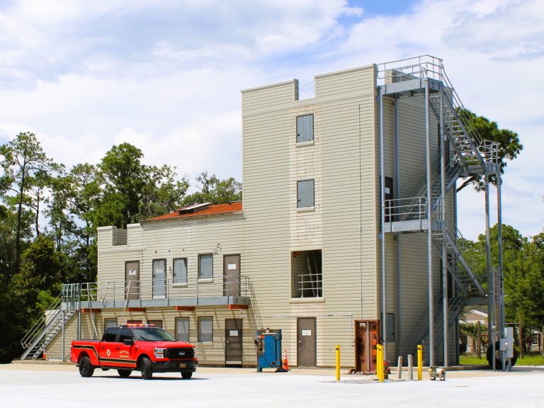 A red fire department pickup truck is parked in front of a multi-story training building with metal stairs and railings. The structure is surrounded by trees under a partly cloudy sky.
