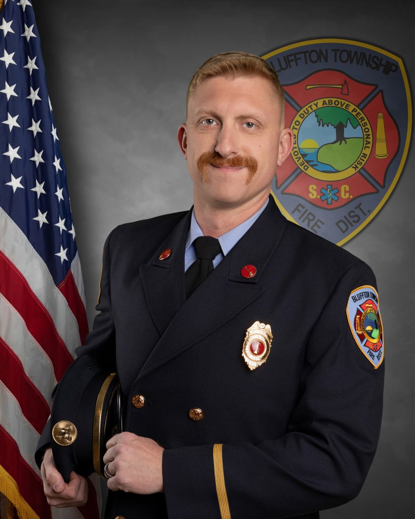 A male firefighter with a blond mustache poses in dress uniform holding a hat, standing in front of an American flag and a Bluffton Township Fire District emblem on a gray backdrop.
