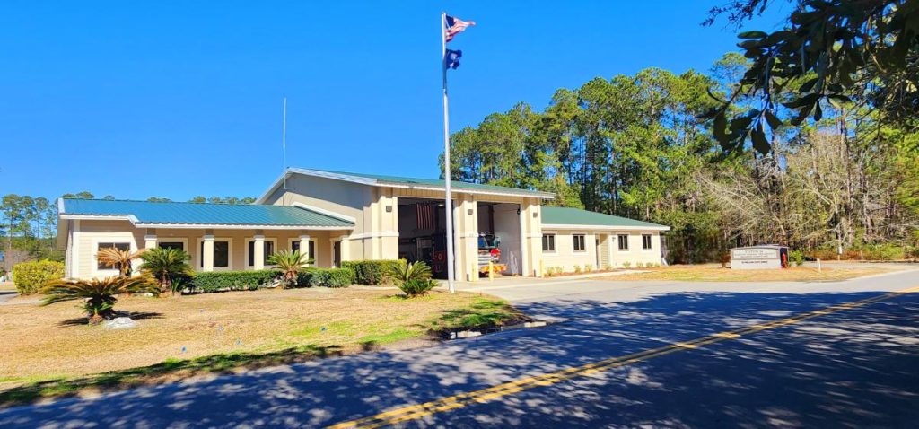 A beige municipal building with a green roof is shown under a clear blue sky. An American flag flies on a pole in front, and a sign is visible near the entrance. Tall trees stand in the background.