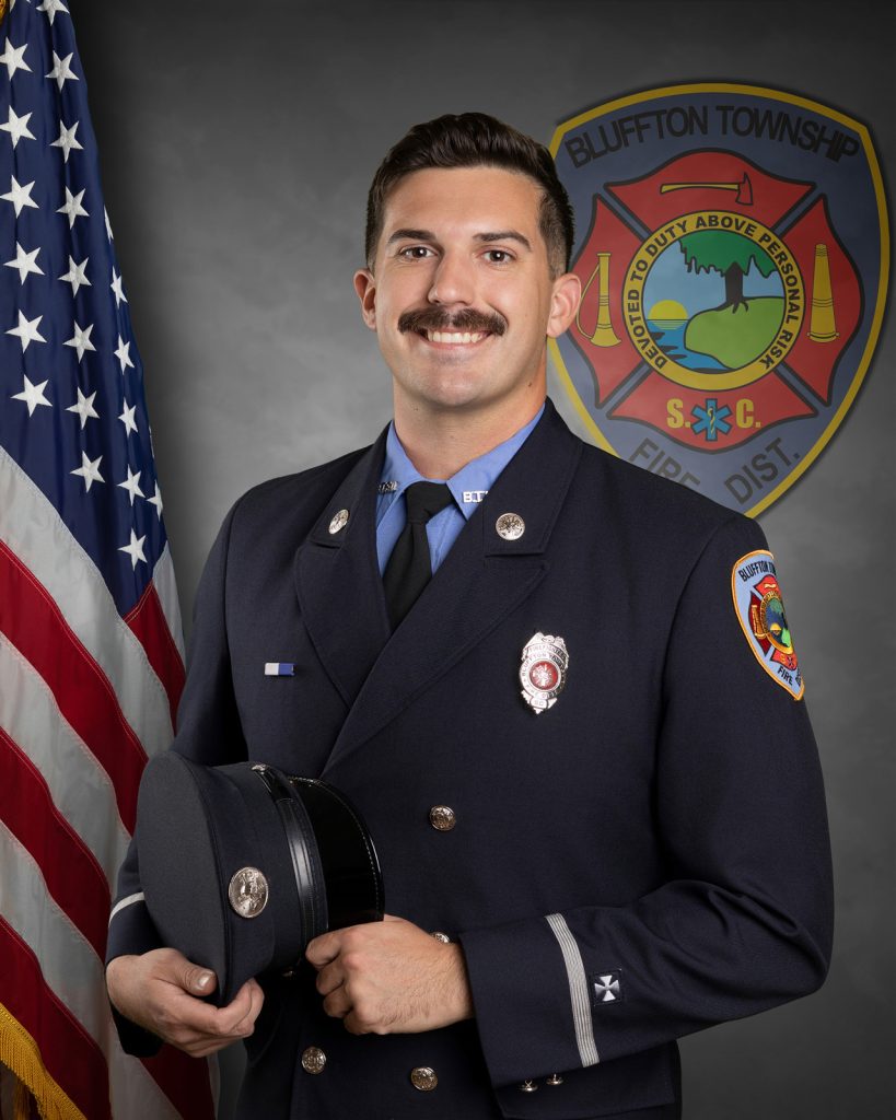 A male firefighter in formal uniform stands holding his hat, smiling, in front of a U.S. flag and the Bluffton Township Fire District emblem on a gray background.