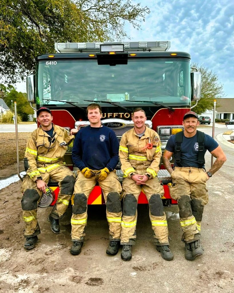 Four firefighters in uniform sit and smile on the front bumper of a red fire truck parked on a residential street, with houses and trees visible in the background under a partly cloudy sky.
