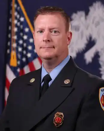 A male firefighter in formal uniform poses in front of an American flag and a blue flag with a white palm tree design. He has short, light brown hair and wears a dark jacket with badges and a patch on the sleeve.