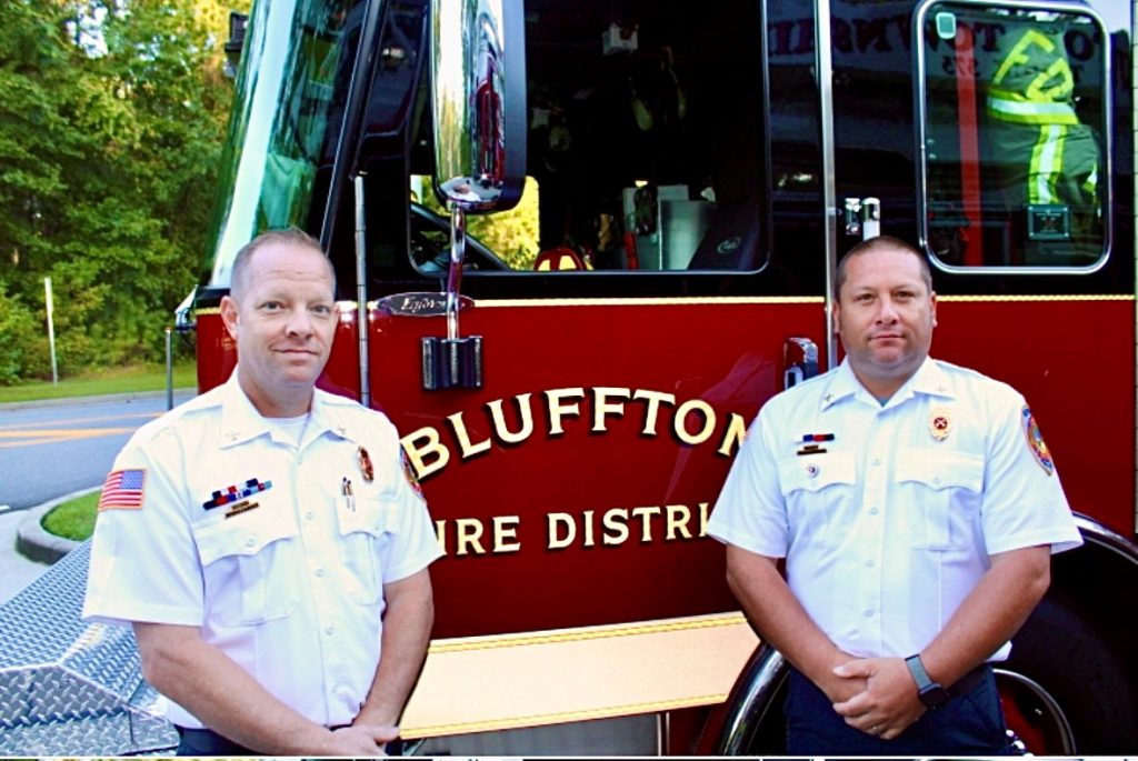 Two uniformed firefighters stand in front of a red Bluffton Fire District fire truck, both looking at the camera, with trees and a road in the background.