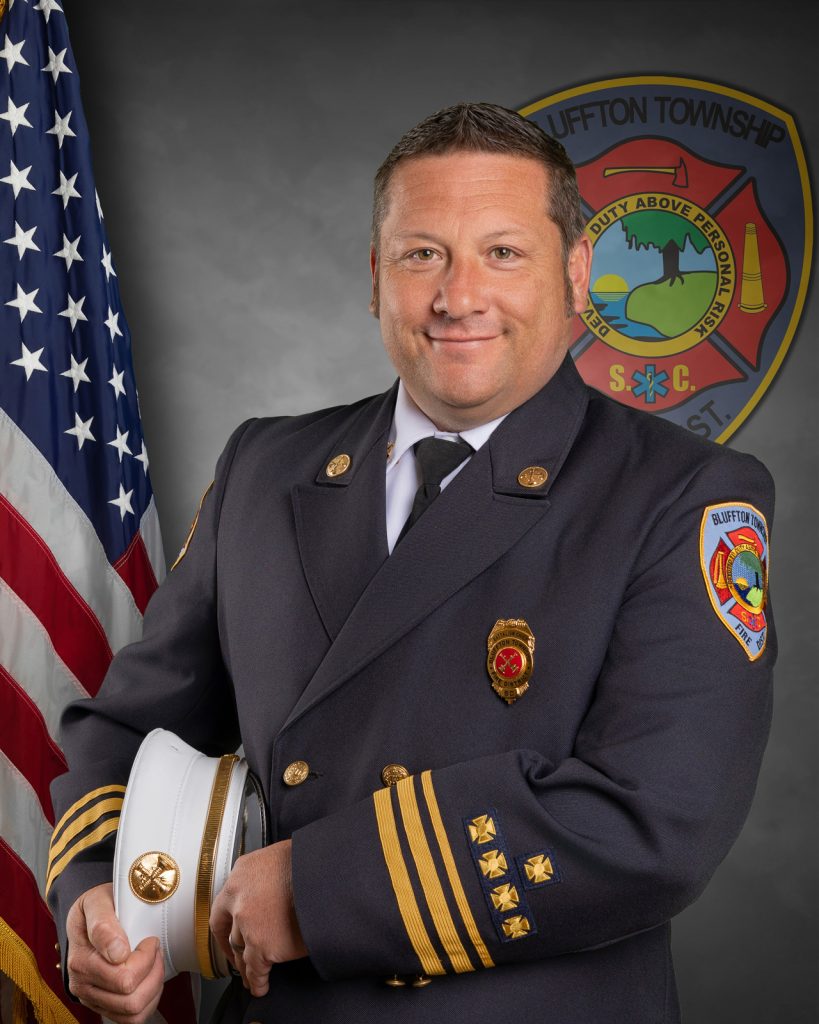 A uniformed fire chief stands in front of an American flag and a Bluffton Township Fire District emblem, holding a white fire chief hat and smiling at the camera.