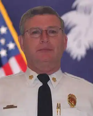 A man in a white uniform with badges and a black tie stands in front of a blurred American flag and another flag featuring a white palmetto tree. He is wearing glasses and looking at the camera.