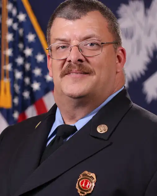 A man wearing a formal dark uniform with a badge and pins poses for a portrait in front of an American flag and another flag in the background.