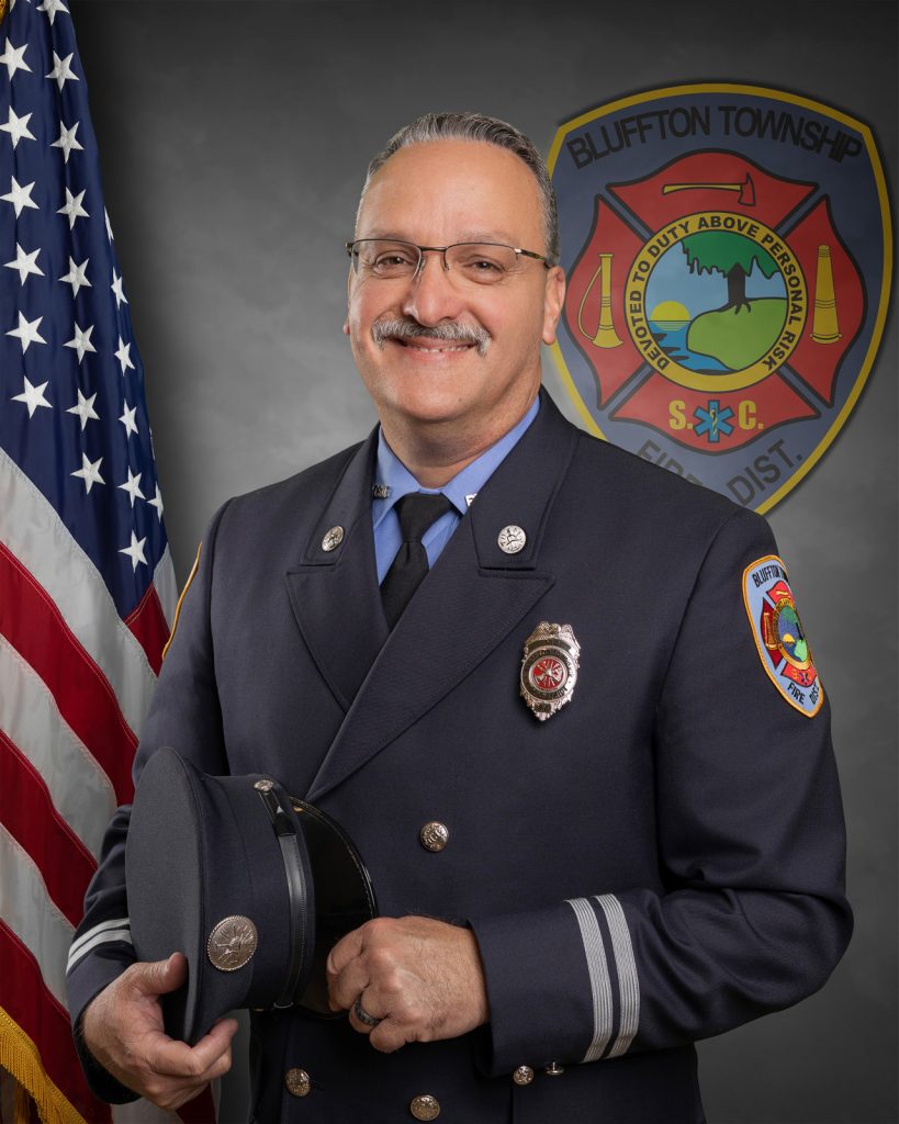 A uniformed firefighter stands smiling in front of an American flag and a Bluffton Township Fire District emblem, holding his cap in his hand.