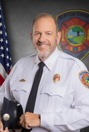 A smiling man in a white fire department dress uniform stands holding a hat. He has a gray beard and wears badges and patches. An American flag and a Bluffton Township Fire District emblem are visible in the background.