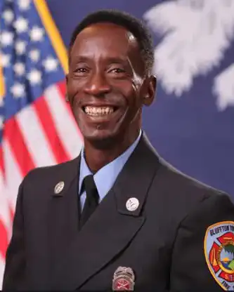 A smiling firefighter in uniform stands in front of an American flag and another flag with white design on blue. A fire department patch is visible on the left sleeve of the uniform.