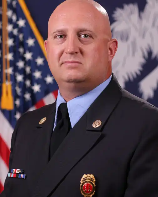 A bald man in a formal dark uniform with badges and a tie stands in front of an American flag and a state flag.