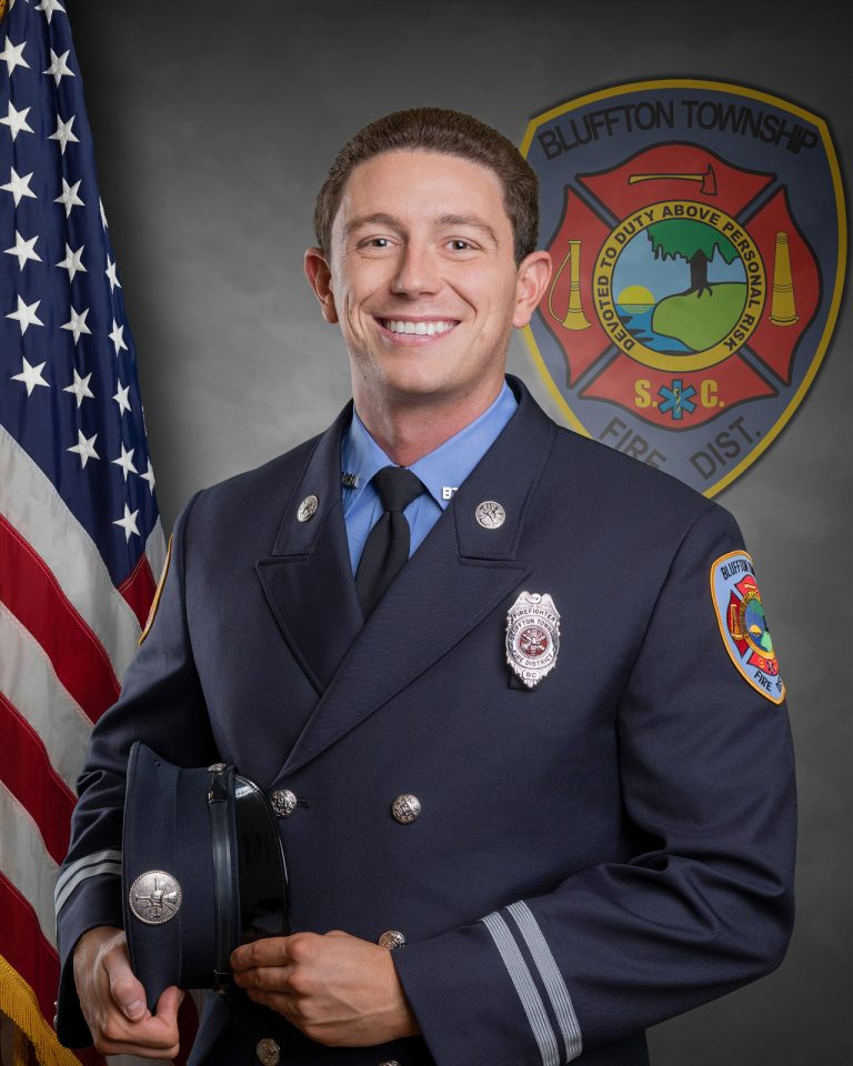 A smiling firefighter in formal uniform holds a hat, standing in front of an American flag and a Bluffton Township Fire District emblem on the wall.