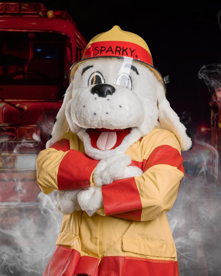 A person in a white dog mascot costume dressed as a firefighter with a yellow helmet labeled "Sparky," stands with arms crossed in front of red fire trucks and smoke.