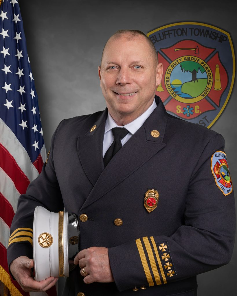 A smiling uniformed fire chief stands in front of an American flag and a Bluffton Township Fire Department emblem, holding his white fire chief's hat under one arm.