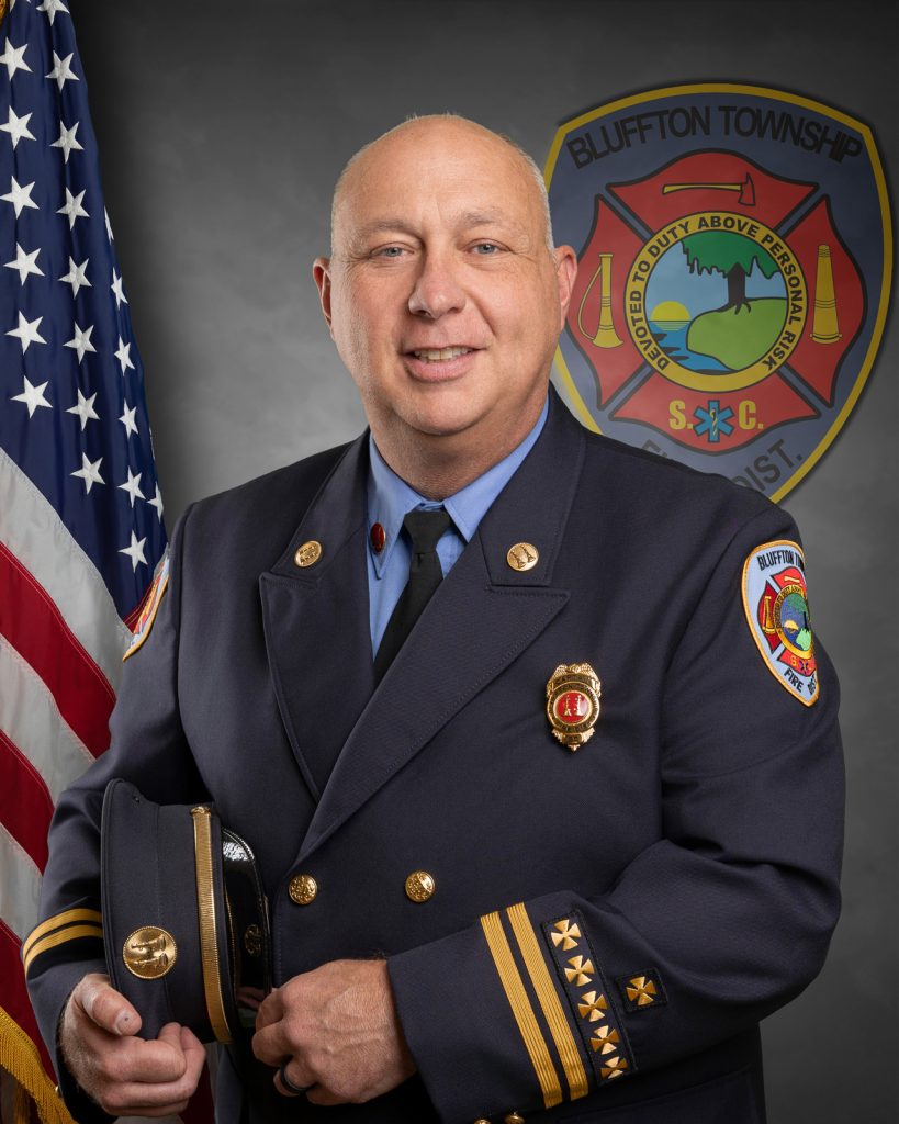 A uniformed fire chief stands smiling in front of an American flag and a Bluffton Township Fire District emblem, holding his hat in his hand.