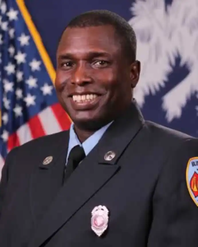 A smiling firefighter in uniform stands in front of the American flag and a blue banner with a white emblem.
