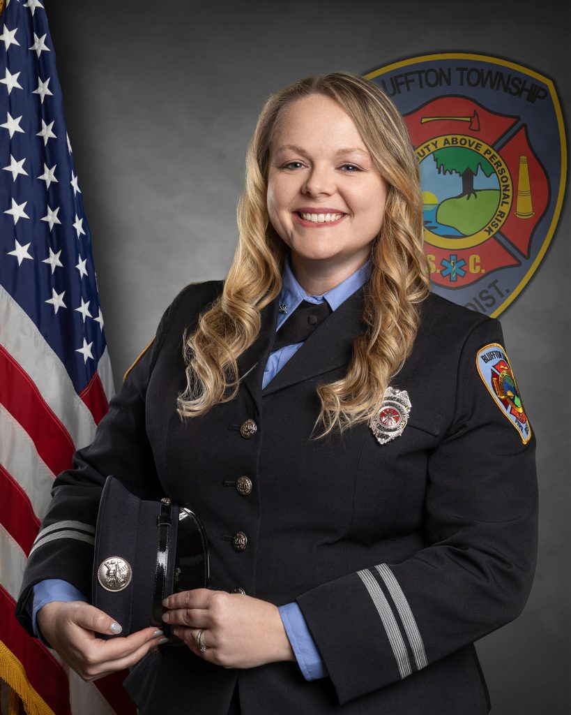 A smiling firefighter in uniform stands holding a hat, posed in front of an American flag and a Bluffton Township Fire District emblem.