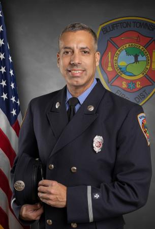 A firefighter in formal uniform stands holding his hat, smiling in front of an American flag and the Bluffton Township Fire District emblem.