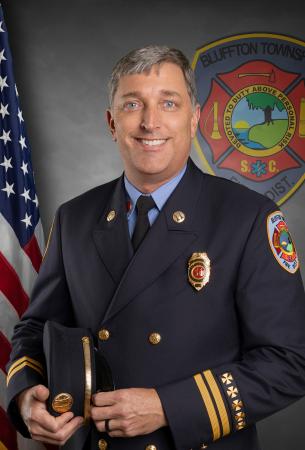 A smiling man in a formal fire department uniform holds his hat. An American flag and a Bluffton Township Fire District emblem are visible in the background.