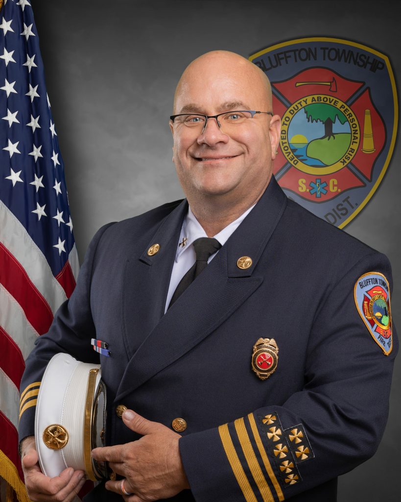 A bald man wearing glasses and a decorated dark firefighter’s uniform stands holding a white hat. An American flag and a Bluffton Township Fire District emblem are visible in the background. He is smiling at the camera.
