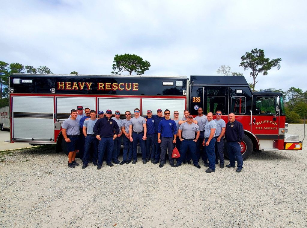 A group of firefighters in uniform pose together in front of a Bluffton Fire District heavy rescue truck parked outdoors on a gravel surface.