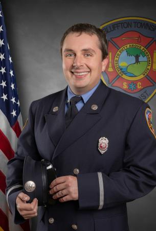 A smiling firefighter in uniform stands holding his hat, with an American flag and a fire department emblem in the background.