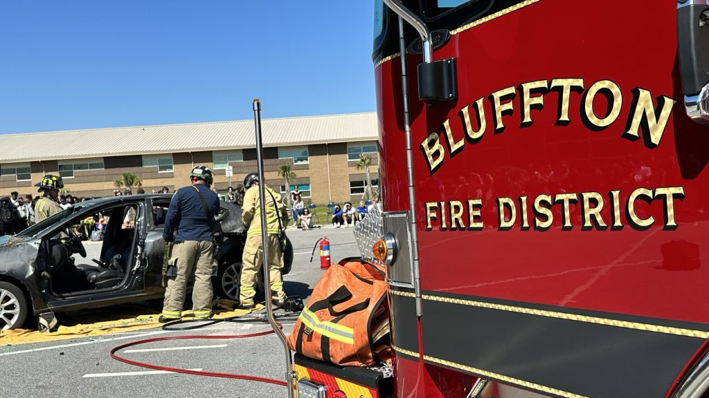 Firefighters work to remove a car door during an emergency demonstration in a parking lot, with a Bluffton Fire District truck in the foreground and a school building in the background.