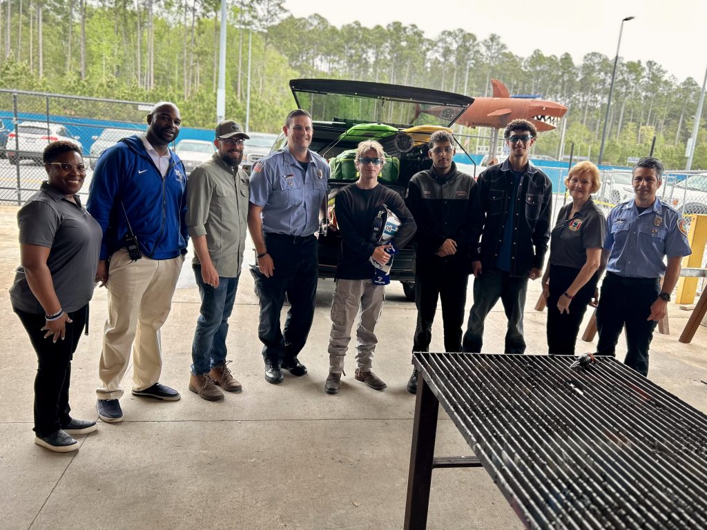 A group of ten people stand together smiling in front of an open car trunk filled with items. They are indoors near a grill, with trees and a fence visible in the background.