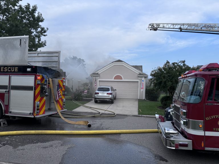 Two fire trucks and hoses are positioned in front of a suburban house with smoke coming from the roof and garage, indicating a fire emergency. A silver car is parked in the driveway.