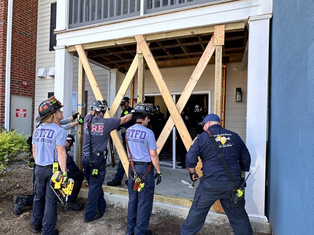 A group of firefighters in uniform stand outside a damaged apartment building entrance, which is supported by large wooden beams arranged in an "X" shape during a rescue or training operation.