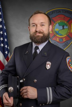 A bearded man in a formal firefighter’s dress uniform holds his hat and stands in front of an American flag and a Bluffton Township Fire District emblem.