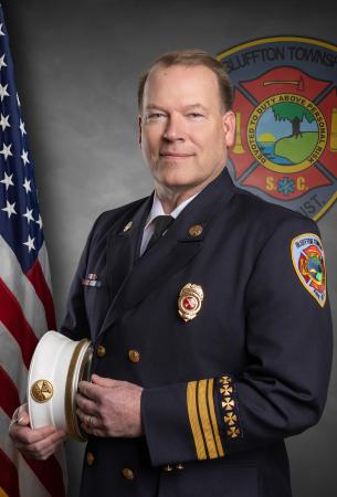 A uniformed fire chief stands holding his white hat, with an American flag and a Bluffton Township Fire District emblem in the background.